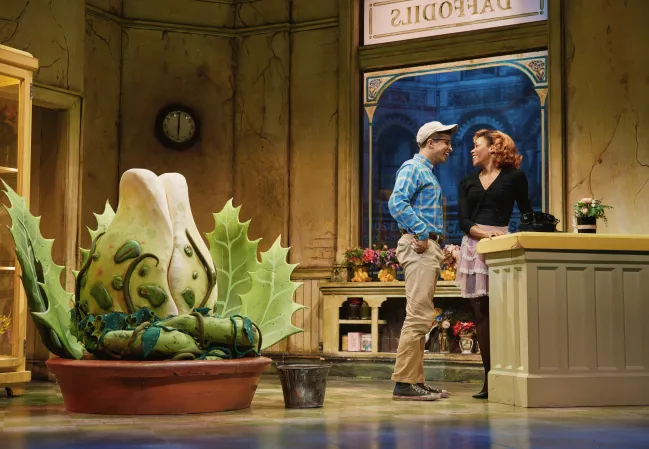 Two people, a man and a woman, standing and talking in an indoor setting with a large plant sculpture and a clock on the wall.
