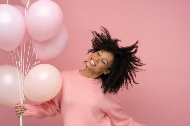 Happy pleased biracial woman with curly hair dancing, holding many balloons, enjoys cool party, wears pink sweater, celebrates birthday, standing over studio pink background