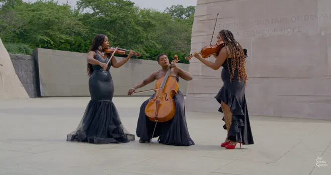 Three musicians performing on a stage, playing violin, cello, and drums, wearing long black dresses.
