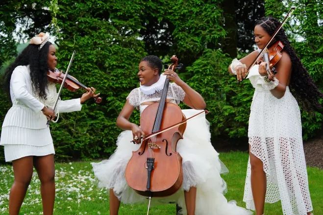 Three women in white dresses playing string instruments in a lush, green outdoor setting.